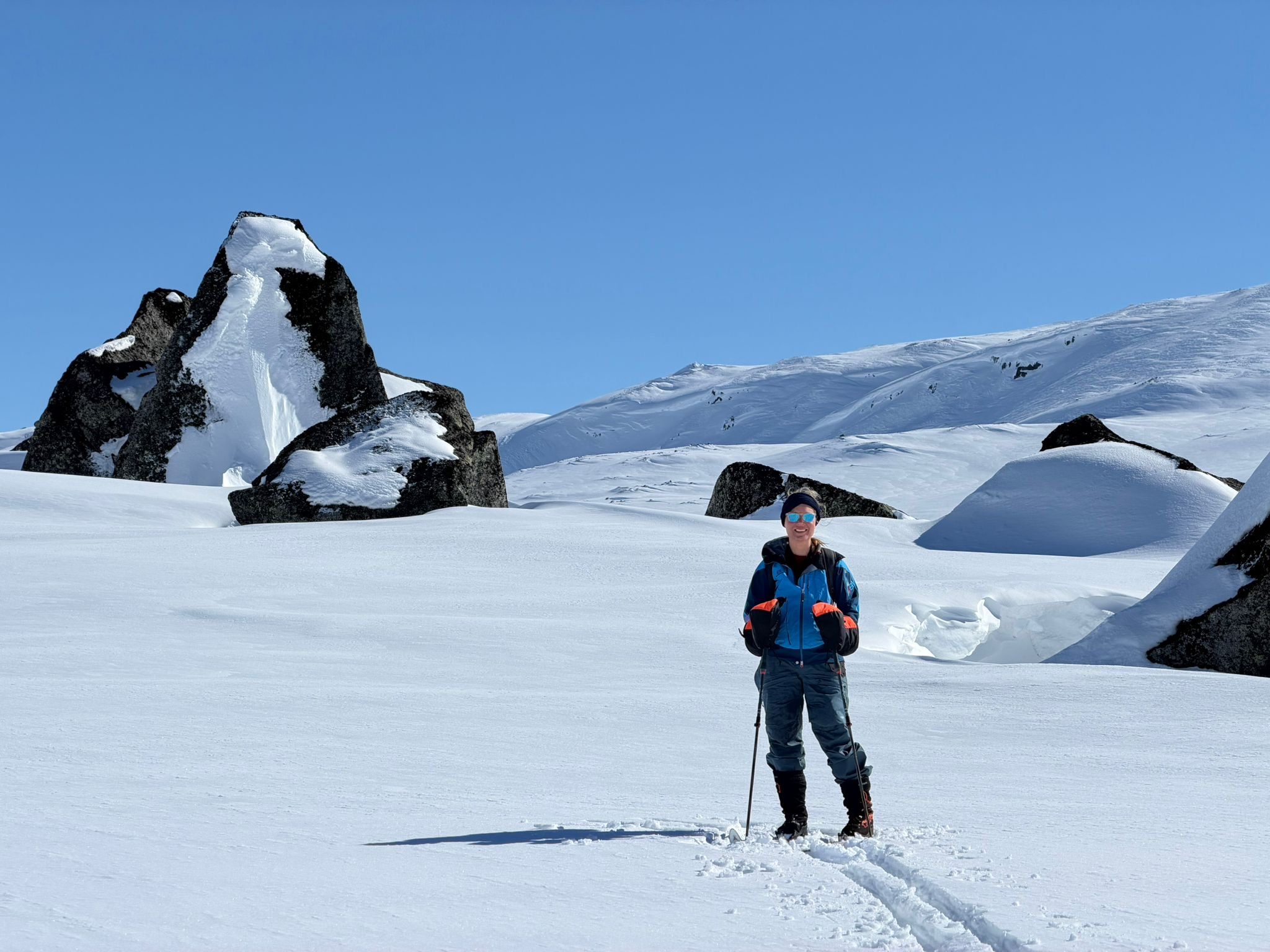 Erica Stanford on skis in a snowy landscape of rock outcrops and rolling snowfields under a clear blue sky