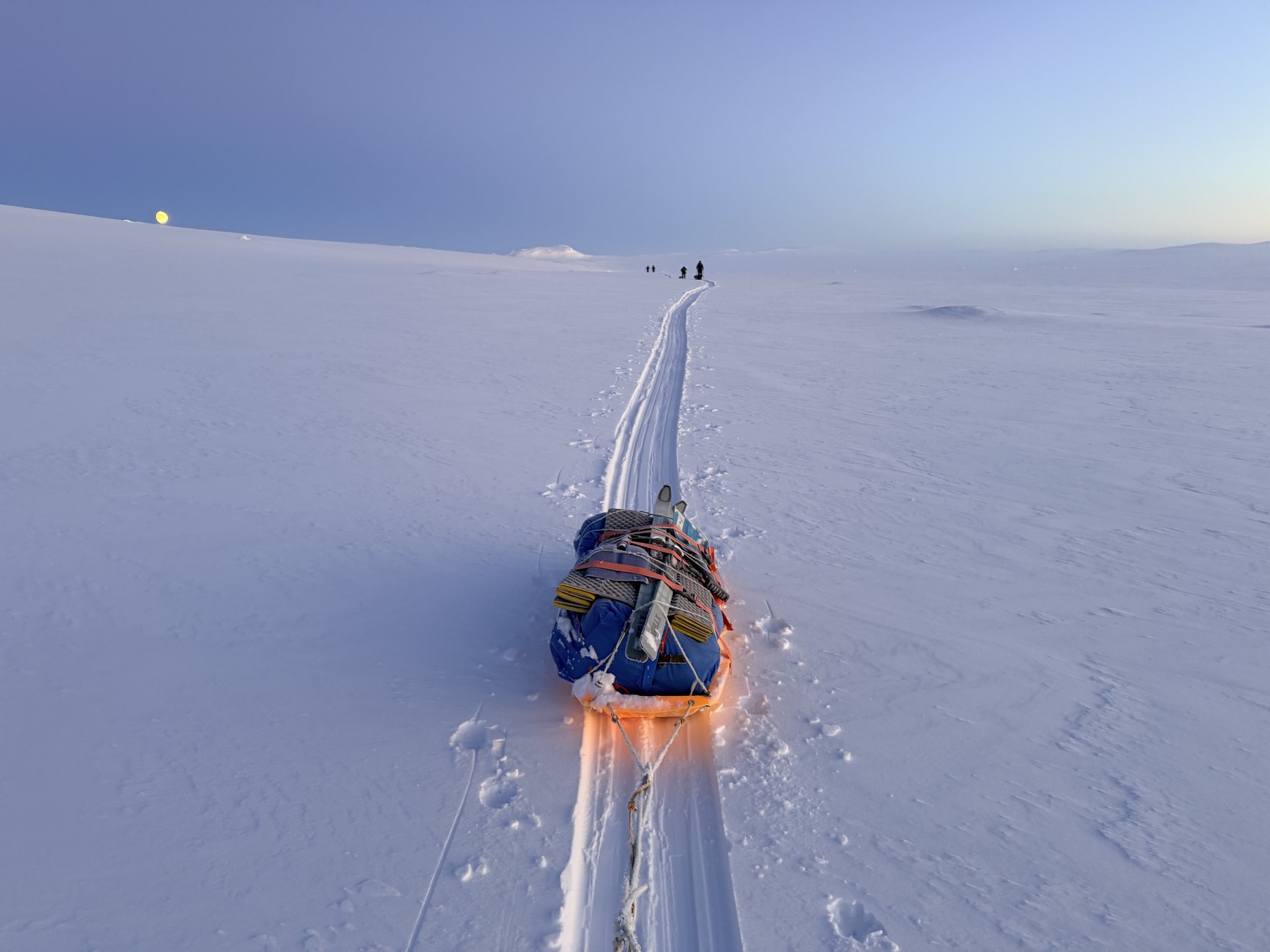 A loaded pulk on the ski track in Halti National Park in Arctic Finland, with the moon low over the snow and the rest of the group ahead on the horizon, during polar training